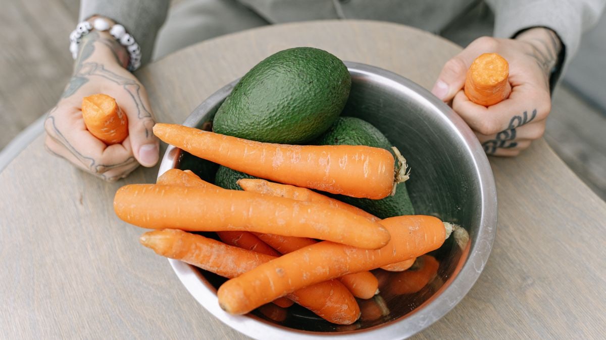 Tattooed person holding a steel bowl filled with fresh avocados and carrots on a wooden table.